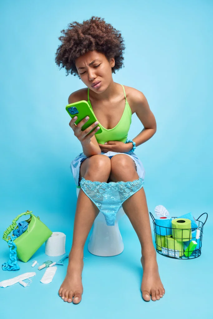 Foto interna de mulher afro-americana sofre de cólicas abdominais tem constipação busca na internet qual remédio tomar segura celular posa no banheiro no vaso sanitário. Problemas de saúde